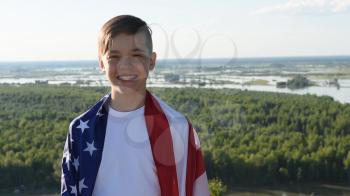 Blonde boy waving national USA flag outdoors over blue sky at the river bank. Beauty summer sunny day. American flag, patriotism, independence day 4th july concept