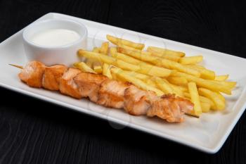 Grilled salmon fish shashlik with fried potatoes and sauce on white plate on black wooden background