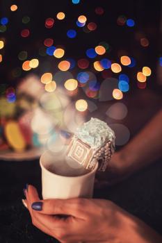 Female hands with cup of tea and christmas cookies house on dark color bokeh lights background