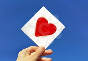Hand holding a picture of a bright red heart against the blue sky