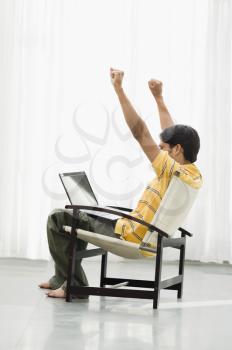 Young man sitting in an armchair with a laptop and his arms raised