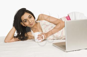 Portrait of a woman lying on the bed with a laptop and holding coffee mug