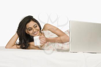 Woman lying on the bed with a laptop and holding coffee mug