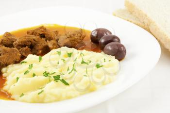 Goulash with mashed potato spiced with parsley, three brown olives and two slices of bread.