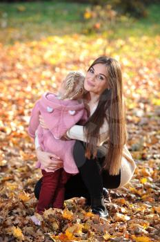A young mother in a sweater hugs her cute blonde daughter during a walk in the autumn Park, against the background of fallen leaves and yellow trees