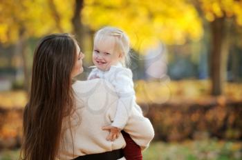 A young mother in a sweater holds her cute blonde daughter in her arms during a walk in the autumn Park, against the background of fallen leaves and trees