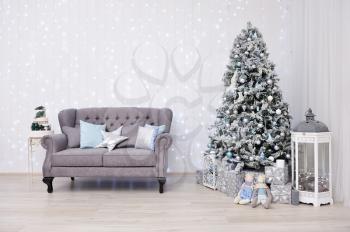 New Year's Decor in a photo studio in gray and white. Classic gray sofa with pillows, snow-white Christmas tree, gifts and a lantern, against the white wall with a garland.