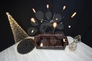 New year's Loft-style decor against a black wall, a Christmas tree from a garland. black chandelier with incandescent lamps and brown sofa. Beautiful New Year's decor with lighting in the studio.