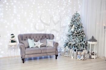 New Year's Decor in a photo studio in gray and white. Classic gray sofa with pillows, snow-white Christmas tree, gifts and a lantern, against the white wall with a garland.