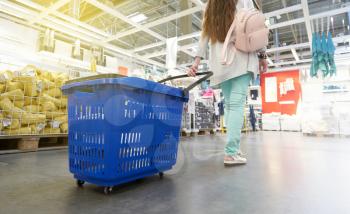 A young girl in bright clothes walks around the store with a blue basket on wheels.