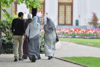 A group of refugees from the Middle East, three women in hijabs and a man walk in the European park and go to the building.