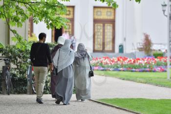 A group of refugees from the Middle East, three women in hijabs and a man walk in the European park and go to the building.