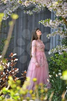 Young girl in a pink fashionable dress, next to a beautiful flowering tree of Japanese cherry in the garden.