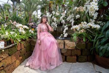 Young girl in a pink fashionable dress among the many orchids in a special winter indoor garden