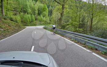 Photo from the window of a car that rides along a road along the forest of Schwarzwald and a cyclist rides nearby. A car rides next to a cyclist