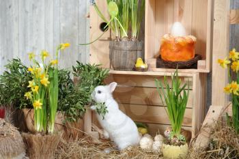 A small and curious white rabbit with blue eyes, stands on its hind legs on dry hay in a studio with Easter decor.