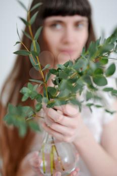 A portrait of a young girl who holds eucalyptus branches inserted into a glass transparent small vase.