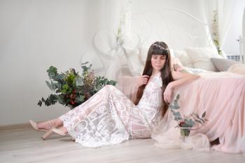 Photo of a young girl in the style of fayn art. A girl in a white translucent dress sits on the floor next to the bed and a bouquet of flowers.