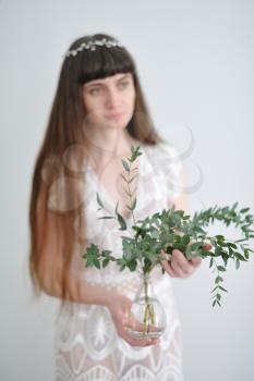 A portrait of a young girl who holds eucalyptus branches inserted into a glass transparent small vase.