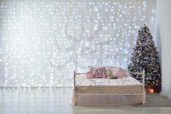 Interior against the background of a white wall with cold garlands, a forged bed and a Christmas tree. Garland on the whole wall, place for text