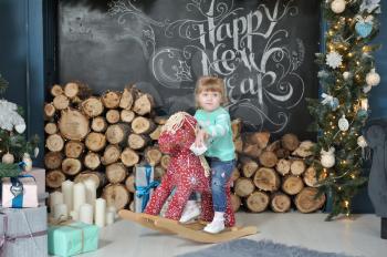 Little girl child sitting on a red rocking horse on the background of the Christmas interior with a fireplace, firewood and gifts