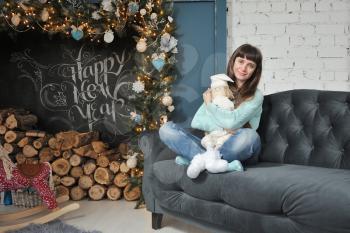 Young girl in jeans sits on a sofa with a soft toy dwarf on the background of Christmas decor and chalk inscription happy new year