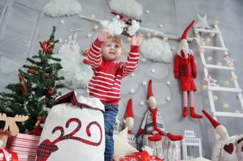 Little girl in a red sweater takes artificial snow from a bag and throws it against the background of the Christmas interior with gnomes, a ladder and garlands