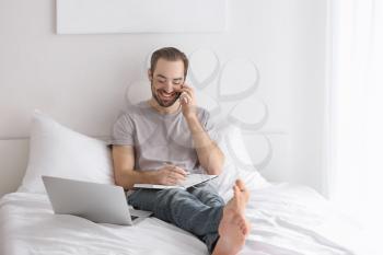 Male freelancer talking on phone while working in bed�