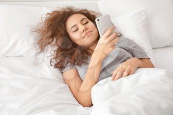 Morning of young African-American woman lying in bed with mobile phone�