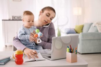 Young mother with baby talking on phone while working at home�