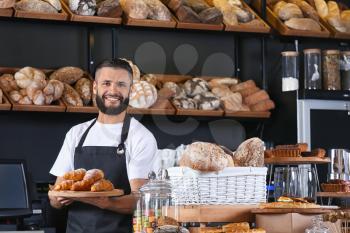Male baker holding wooden board with delicious croissants in shop�
