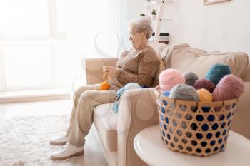 Senior woman sitting on sofa while knitting sweater at home�