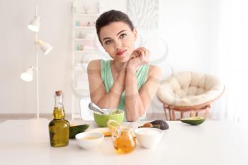 Young woman making nourishing mask with avocado in kitchen�