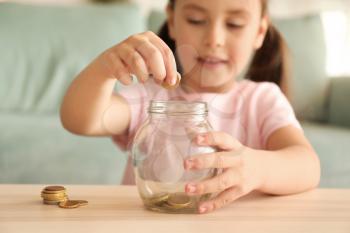 Little girl putting coins into glass jar indoors. Money savings concept�