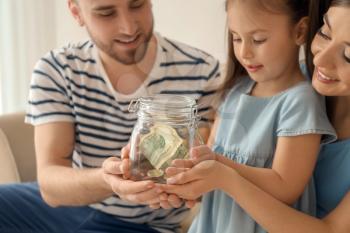 Little girl with her parents holding glass jar with dollars indoors. Money savings concept�