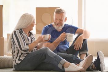 Mature couple with belongings sitting on carpet and drinking tea indoors. Moving into new house�