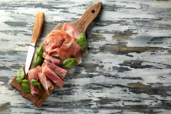 Wooden board with tasty prosciutto slices and basil on table�