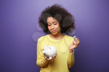Young African-American woman with money and piggy bank on color background. Savings concept�