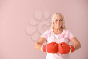 Beautiful mature woman with pink ribbon and boxing gloves on color background. Breast cancer concept�