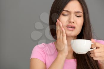 Young woman with sensitive teeth and cup of hot coffee on grey background�