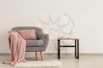 Comfortable armchair with table and lamp near light wall�
