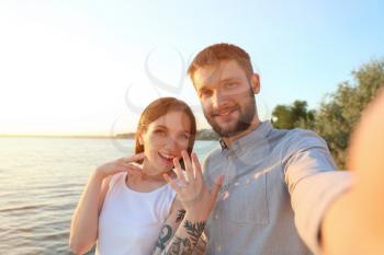 Happy couple taking selfie with engagement ring near river on sunny day�