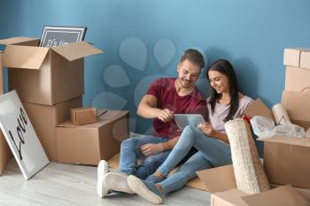 Young happy couple with tablet PC and moving boxes sitting on floor at new home�
