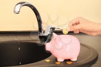 Woman putting coin into piggy bank near metal tap. Water saving concept�