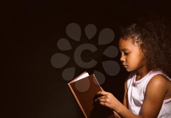 Little African-American girl reading book on dark background�