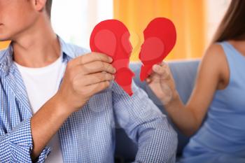 Young couple holding halves of broken heart on sofa at home. Relationship problems�