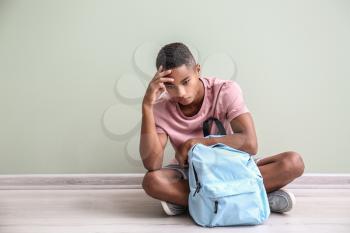 Sad African-American teenage boy sitting on floor indoors. Bullying at school�