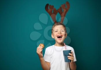 Cute little boy with toy reindeer horns holding cup of hot chocolate and cookie on color background�