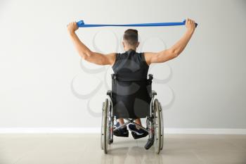 Sporty man training with elastic band while sitting in wheelchair against light wall�