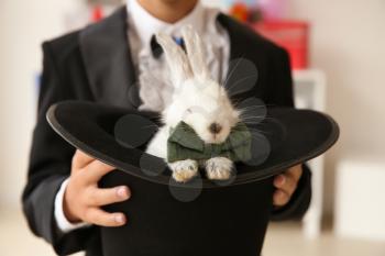 Cute little magician holding hat with rabbit indoors, closeup�